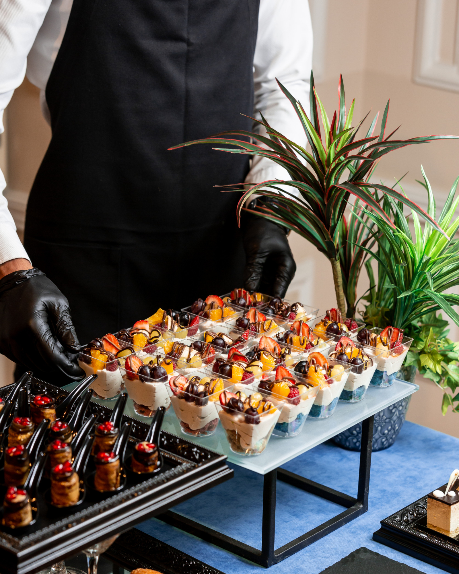 Personale di servizio con guanti neri che dispone bicchierini di dessert monoporzione alla frutta e dolci mignon su un tavolo del buffet curato da un catering per matrimoni.