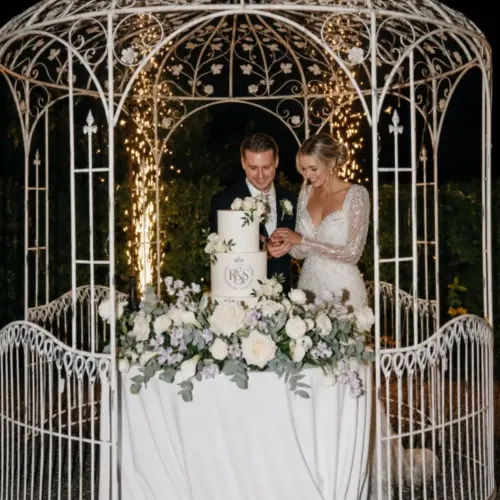 Sposi durante il taglio della torta nuziale con catering per matrimoni in una raffinata location all'aperto illuminata da luci decorative.