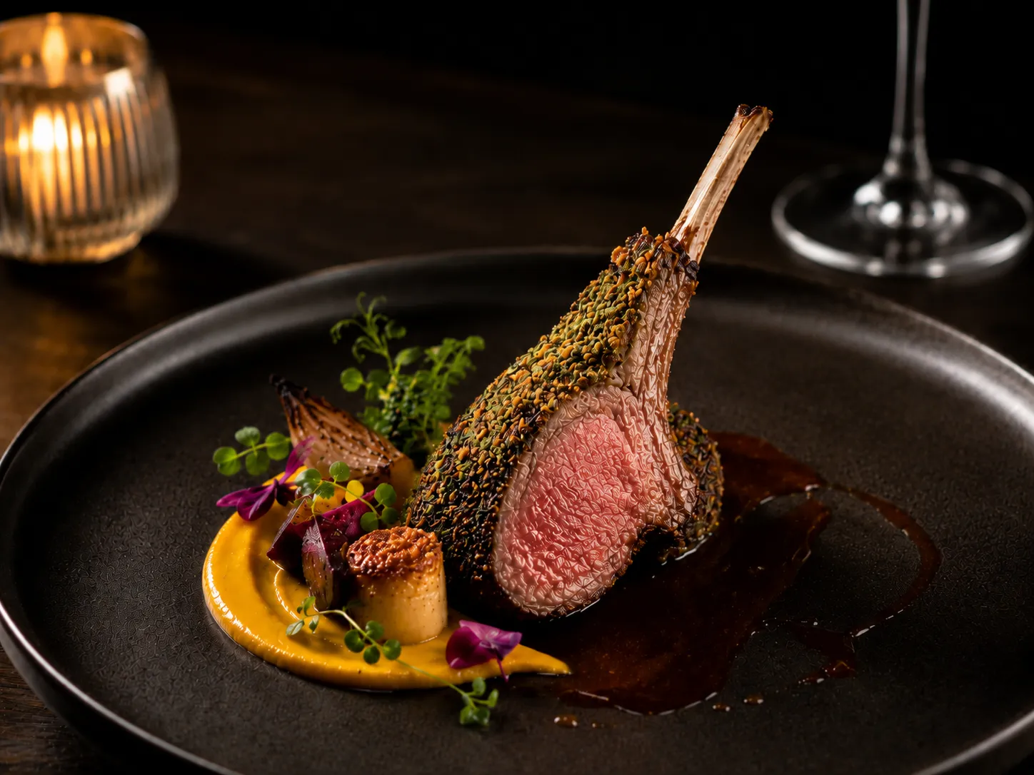 Plated rack of lamb with sauce, vegetable purée, herbs and garnish served on a black plate